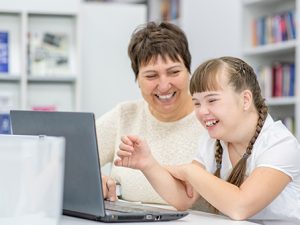 A support teacher laughs with a young pupil she is assisting whilst they work on a laptop