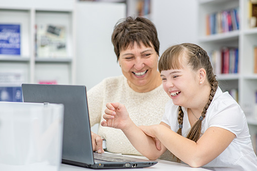 A support teacher laughs with a young pupil she is assisting whilst they work on a laptop